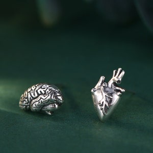 May include: A pair of silver earrings, one shaped like a brain and the other shaped like a heart. The earrings are on a green background.