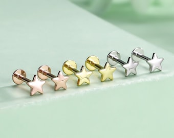 May include: A set of six star-shaped stud earrings in different metal finishes. The earrings are arranged in a row on a light green surface. The finishes include silver, gold, and rose gold.