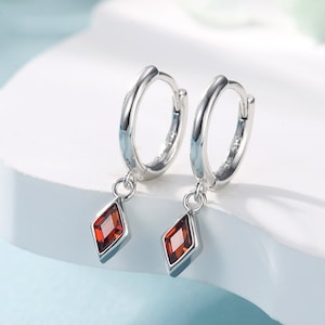 Puede incluir: Un par de pendientes de aro de plata con una piedra preciosa roja en forma de diamante.