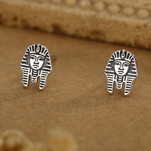 May include: A pair of silver stud earrings depicting the face of an Egyptian pharaoh. The earrings have a detailed design with a headdress and facial features. The earrings are presented on a brown surface.