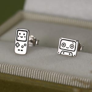 Puede incluir: Pendientes de plata con diseño de consola de videojuegos retro y cinta de cassette. Los pendientes son pequeños, con un contorno detallado de cada forma. Los pendientes se muestran en una caja de joyería de terciopelo verde.