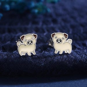 May include: A pair of silver pug dog stud earrings. Each earring depicts a cream-coloured pug with brown accents on the ears and a black eye. The earrings are set against a dark blue textured background.