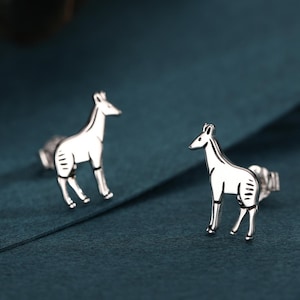 May include: Two silver-colored stud earrings, each shaped like a giraffe, are displayed on a dark teal surface. The giraffes have detailed legs, bodies, and necks. The earrings are small and delicate, with a polished finish.