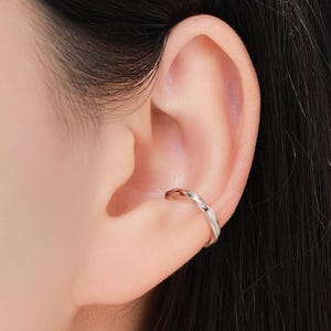 May include: A silver ear cuff with a twisted design is worn on the ear. The jewelry is a simple, elegant accessory. The ear cuff is a small, delicate piece of jewelry.