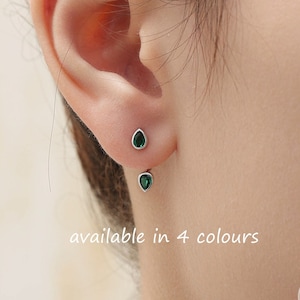 Puede incluir: Un par de pendientes de plata con pequeñas piedras verdes en forma de lágrima. Los pendientes se llevan en el lóbulo superior de la oreja. El texto "available in 4 colours" está escrito debajo de los pendientes.