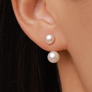 May include: A double-sided earring featuring two white spherical pearls of different sizes. The smaller pearl sits directly on the earlobe, while the larger pearl is positioned below the lobe, creating a layered and balanced aesthetic appearance.