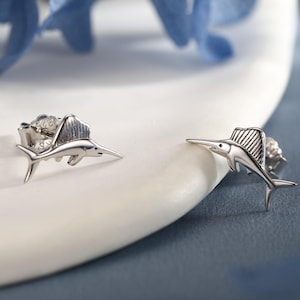 Puede incluir: Un par de pendientes de plata con diseño de pez vela. Los pendientes presentan detalles de aletas y un hocico largo y puntiagudo. Se encuentran sobre una superficie blanca, con acentos florales azules en el fondo.