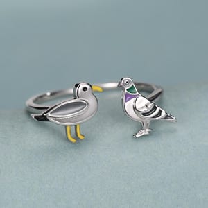 May include: A silver-toned open ring featuring two bird figures facing each other. One bird is a white and grey seagull with a yellow beak and feet. The other bird is a grey pigeon with a purple and green neck accent. The band is thin and metallic.