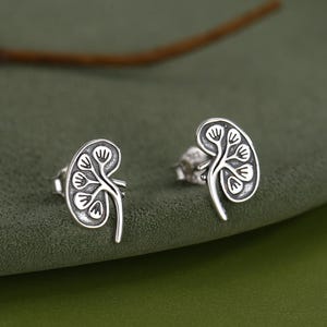 Op de afbeelding: Een paar zilverkleurige oorstekers in de vorm van een nier. De oorbellen hebben een gedetailleerd ontwerp met een boomachtig patroon in de nier. De oorbellen worden gepresenteerd op een groene ondergrond.