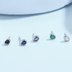 May include: Five teardrop-shaped earrings with different coloured gemstones. The earrings are silver and have a small post for piercing. The gemstones are black, blue, purple, green, and white.