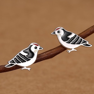 May include: A pair of stud earrings shaped like small birds. Each bird features a white body, black wings with white stripes, a black beak, and a small red accent on the top of the head. The earrings are crafted with smooth enamel detailing.