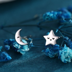 Könnte beinhalten: Ein Paar silberne Ohrringe mit einem Halbmond und einem Stern mit einem lächelnden Gesicht. Die Ohrringe befinden sich auf einem blauen Hintergrund mit blauen Blumen.