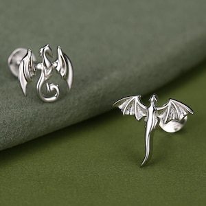 May include: Two silver stud earrings featuring dragon designs. One earring depicts a curled dragon with folded wings, while the other shows a dragon with outstretched wings and a long tail. Both pieces have a polished metallic finish and post backs.