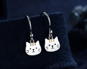 May include: A pair of silver cat-shaped dangle earrings with white enamel and gold accents. The earrings are displayed on a dark blue velvet background.