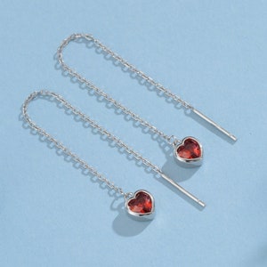 Pode incluir: Um par de brincos de prata em forma de coração com detalhes de pedras preciosas vermelhas pendurados em correntes delicadas.