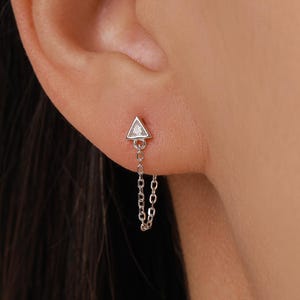 May include: A silver earring with a small triangle-shaped top and a dangling chain. The earring is worn on the ear, showcasing its delicate design and minimalist aesthetic. The chain adds movement and visual interest to the piece.