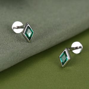 Könnte beinhalten: Ein Paar silberne Ohrstecker mit einem rautenförmigen, smaragdgrünen Edelstein. Die Ohrringe sind vor einem grünen Hintergrund platziert, mit einer unscharfen Pflanze oben links. Die Ohrringe haben einen Stecker und einen Verschluss.