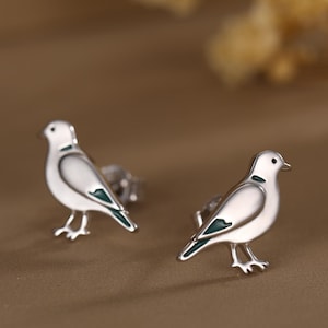 May include: A pair of silver-colored dove stud earrings. Each earring features a white body with a teal accent on the wing and neck. The earrings are set against a brown background with a blurred floral element.