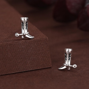Peut inclure: Une paire de boucles d'oreilles clous argentées en forme de bottes de cowboy. Chaque botte présente des détails noirs complexes et un petit accent scintillant. Les boucles d'oreilles sont présentées sur une surface marron.