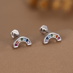 May include: A pair of silver rainbow stud earrings. Each earring has a curved rainbow design, set with a variety of coloured gemstones. The gemstones include red, orange, yellow, green, blue, and purple. The earrings have a post and backing for secure wear.