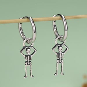 Può includere: Un paio di orecchini a cerchio in argento con ciondoli a forma di scheletro. Ogni orecchino è composto da un cerchio lucido con una piccola figura scheletrica dettagliata appesa, con le braccia sollevate sopra la testa in posizione fissa.
