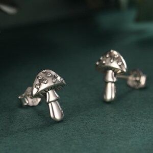 May include: A pair of silver mushroom stud earrings. The mushroom caps are dotted with small, clear stones. The earrings are set against a dark green background, with a blurred leaf in the upper left corner.