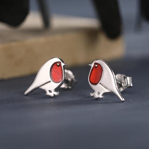 Può includere: Un paio di orecchini a lobo argentati a forma di pettirossi. Ogni uccello ha il petto rosso e si staglia su uno sfondo scuro. Gli orecchini sono piccoli e dettagliati, con un design ispirato alla natura.
