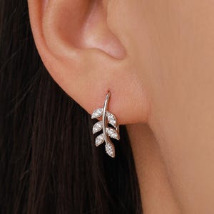 Può includere: Un orecchino a forma di foglia d'argento con piccole gemme trasparenti. L'orecchino ha un design delicato, con un gancio curvo e un motivo a foglie dettagliato. Le gemme aggiungono brillantezza.
