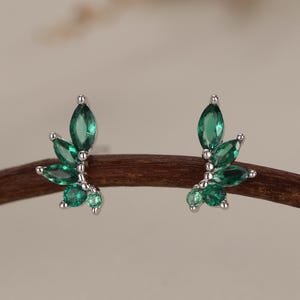 May include: A pair of silver stud earrings featuring a leaf design. Each earring has multiple marquise-cut, emerald-green gemstones arranged to resemble leaves. The earrings are displayed on a brown branch against a neutral background.