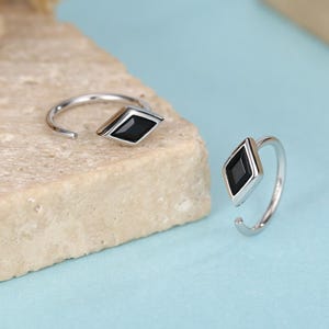Puede incluir: Dos pendientes de aro plateados con una piedra negra en forma de diamante. Los pendientes están abiertos en la parte inferior y descansan sobre una superficie de piedra clara sobre un fondo azul. Flores secas en el fondo.