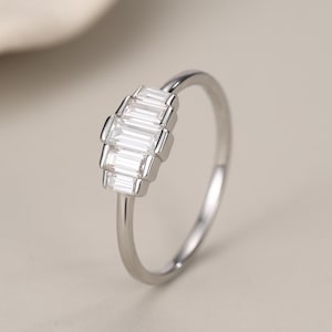May include: A silver-toned ring featuring a cluster of five clear, rectangular baguette-cut stones arranged in a stepped, fan-like formation. The band is thin and polished, showcasing a metallic finish that complements the clear, faceted stones.