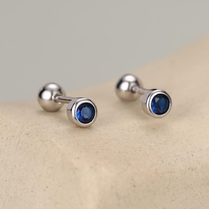 Peut inclure: Une paire de boucles d'oreilles à tige de couleur argentée avec une pierre précieuse bleue ronde sertie clos. Chaque boucle d'oreille possède une tige en métal poli lisse et un fermoir sphérique, offrant un design épuré et simple.
