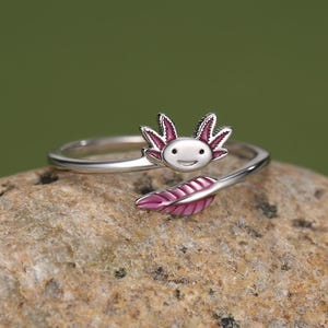 Könnte beinhalten: Ein silberfarbener offener Ring mit einem stilisierten Axolotl-Kopf mit rosa akzentuierten Kiemen an einem Ende und einem geschwungenen, rosa schattierten blattartigen Schwanz am anderen Ende, der ein umlaufendes Design bildet.