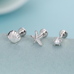 May include: Three silver-colored stud earrings with ocean-themed designs. One earring features a seashell, another a starfish, and the third a small pearl. The earrings are displayed on a light blue surface against a blue background.