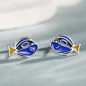 May include: A pair of stud earrings featuring a blue fish design with black and yellow accents. The earrings have a silver-colored metal finish and a smiling face. The fish design is a popular cartoon character.
