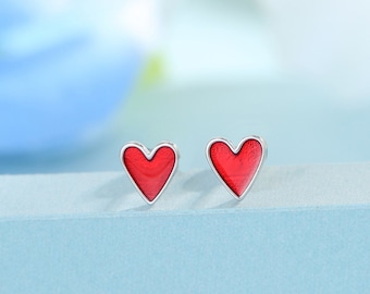 Puede incluir: Un par de pendientes de plata en forma de corazón con esmalte rojo. Los pendientes son pequeños y delicados.