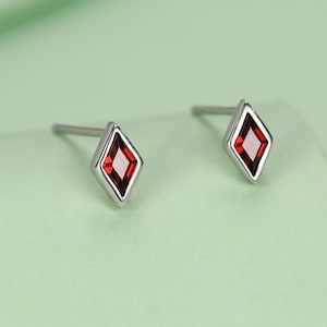 May include: A pair of silver stud earrings. Each earring has a red, diamond-shaped gemstone set in a silver frame. The earrings are shown on a light green surface, with a soft focus background.