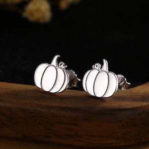 May include: A pair of silver pumpkin stud earrings with white enamel detailing. The earrings are shaped like pumpkins, with a stem and curved lines. They are displayed on a wooden surface, with a dark background.