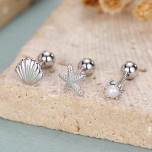 May include: Silver-toned stud earrings featuring seashell, starfish, and pearl designs. The earrings are displayed on a textured, light-colored surface with dried flowers in the background. The earrings have a screw-back closure.