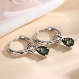 May include: A pair of silver hoop earrings with teardrop-shaped green and black gemstones. The earrings are displayed on a cream-colored surface with a soft, blurred background featuring pink flowers.