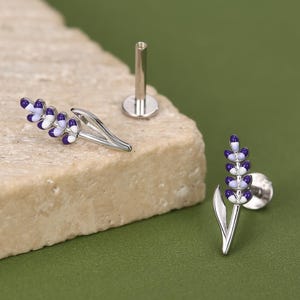 May include: A pair of silver-toned stud earrings shaped like lavender sprigs, featuring purple and white enamel detailing on the flower buds. One earring is shown from the side, while the other displays the flat back post and circular backing.