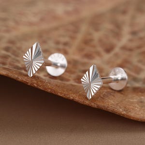 May include: A pair of silver-toned stud earrings featuring a diamond-shaped front face with a sunburst-style engraved pattern. Each earring has a straight post and a flat, circular backing, designed for secure placement in a piercing.