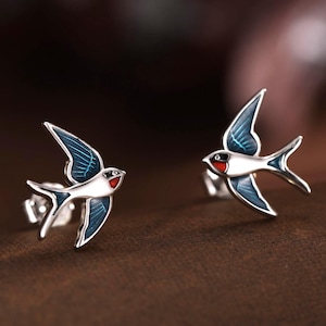 May include: A pair of silver swallow stud earrings. Each earring features a bird with white and blue wings and a red breast. The earrings are presented on a brown surface.
