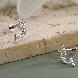 May include: Two silver mermaid-shaped ear studs. The mermaid design features a detailed head and tail. The studs are displayed on a textured, light-colored surface against a soft green background. The earrings are small and delicate.