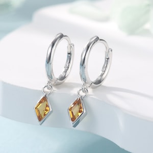 May include: A pair of silver hoop earrings with a dangling diamond-shaped gemstone. The gemstone is a light amber color.