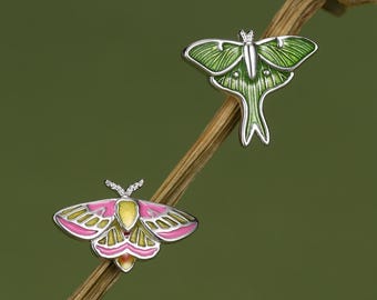 Könnte beinhalten: Zwei Metall-Emaille-Anstecknadeln in Form von Motten. Eine Nadel zeigt eine grüne Motte mit silbernen Umrissen und einem langen Schwanz. Die zweite Nadel stellt eine Motte mit rosa und gelben Flügeln dar, ebenfalls mit silbernen Umrissen.