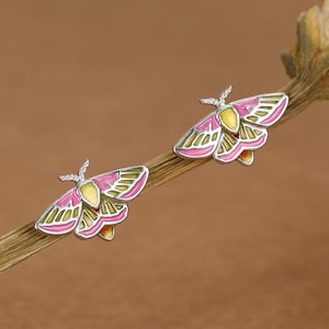 May include: A pair of silver stud earrings shaped like moths, featuring enamel detailing in vibrant shades of pink and yellow. The moth wings display a symmetrical pattern with pink outer edges and yellow centres, finished with silver antennae.