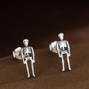 May include: A pair of silver skeleton stud earrings. Each earring features a detailed miniature skeleton design. The earrings are displayed on a brown surface, with a blurred brown background. The earrings are a unique accessory.