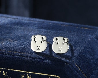Puede incluir: Un par de pendientes de plata con un pequeño diseño de conejillo de indias detallado. Los pendientes se muestran sobre un fondo de terciopelo azul.