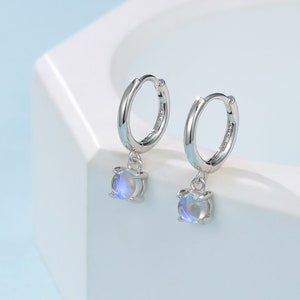 Puede incluir: Un par de pendientes de aro de plata con una pequeña piedra preciosa redonda e iridiscente que cuelga de cada aro.